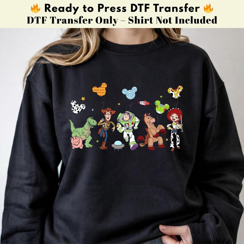 Pode incluir: Uma camisola preta de manga comprida com uma impress&atilde;o gr&aacute;fica de personagens animados, incluindo um cowboy, um ranger espacial, uma cowgirl, um dinossauro, um porco e um cavalo. O design inclui bal&otilde;es coloridos em forma de orelhas.