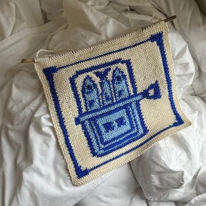 May include: A square, off-white crochet wall hanging with a blue geometric design. The design features a central square with a stylized building motif in shades of blue, framed by a blue border. A wooden dowel is attached at the top.