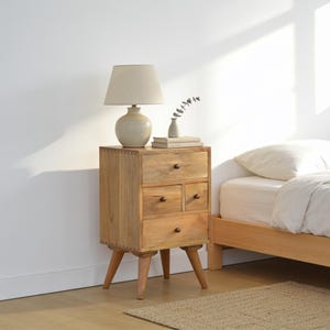 May include: A light brown wooden bedside table with four drawers, featuring visible dovetail joints on the corners and four tapered legs. A cream-coloured table lamp with a rounded base and a small white vase with a sprig sit on the top surface.