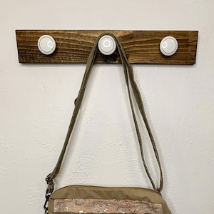 Puede incluir: Un bolso bandolera de lona verde con cierre de cremallera y un gran bolsillo de parche en la parte delantera. El bolsillo tiene un diseño floral rojo. El bolso tiene una correa marrón y está colgado de un estante de madera.