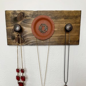 Pode incluir: Um organizador de joias de madeira com três ganchos e uma pequena tigela de terracota. Os ganchos são adornados com botões pretos. Três colares pendurados nos ganchos, um com pingentes de pedras preciosas vermelhas, um com um pingente de ouro que diz "FEMINIST AF", e um com um pingente de pedras preciosas vermelhas.