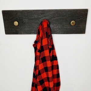 Pode incluir: Um cabide rústico de madeira escura com dois botões de latão. Uma camisa de flanela xadrez vermelha e preta está pendurada no cabide. O cabide está montado em uma parede branca.