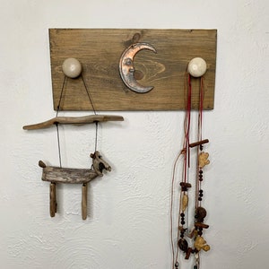 Pode incluir: Um cabide de parede de madeira com duas maçanetas de cerâmica branca e um design de lua crescente de cobre. Uma escultura de madeira de um cavalo está pendurada em uma maçaneta e um colar de contas está pendurado na outra.