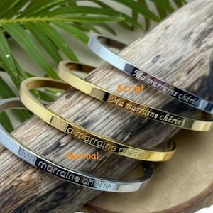 Peut inclure: Quatre bracelets en métal, deux argentés et deux dorés, sont présentés sur un morceau de bois. Chaque bracelet est gravé de la phrase "Ma marraine chérie". Le texte est en écriture script et normale.