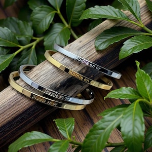 Peut inclure: Quatre bracelets en métal, deux argentés et deux dorés, sont disposés sur une surface en bois avec des feuilles vertes. Chaque bracelet porte l'inscription "Mamie en or".