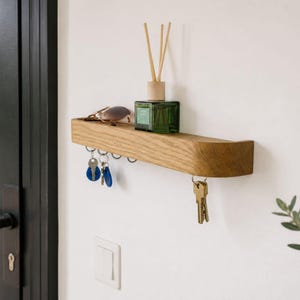 Peut inclure: Une étagère murale en bois clair dotée d'une série de crochets métalliques sur le bord inférieur pour suspendre des clés. Un flacon en verre vert avec un bouchon en bois et des bâtonnets diffuseurs est posé dessus, avec des lunettes de soleil.