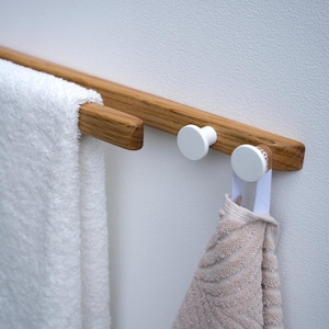 Puede incluir: Toallero de madera con acabado natural, con una barra para colgar toallas y dos pomos blancos. Una toalla blanca cuelga a la izquierda, y una toalla beige está suspendida de uno de los pomos. Montado en una pared blanca.