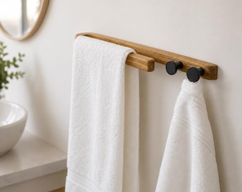 May include: A wall-mounted towel rack featuring a horizontal light brown wooden bar with two black circular hooks and a separate wooden arm for hanging towels. Two white cotton towels are draped over the rack, one on the arm and one on a hook.