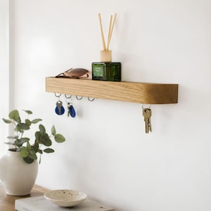 May include: A light-colored wooden wall-mounted shelf featuring a flat top surface and a row of metal hooks underneath. The shelf holds a green glass bottle with reed diffuser sticks and a pair of sunglasses. Several sets of keys hang from the hooks.
