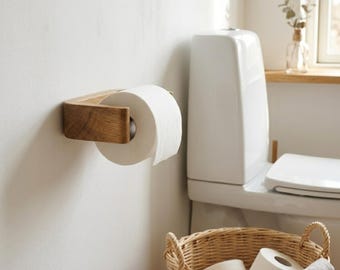 May include: A wall-mounted toilet paper holder crafted from light brown wood with a visible grain, holding a roll of white toilet paper. Below, a woven wicker basket contains additional rolls of white toilet paper and a folded beige fabric cloth.