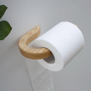 Puede incluir: Un portarrollos de papel higiénico de madera con un diseño curvo. El soporte es de madera de color claro y sostiene un rollo de papel higiénico blanco. El papel tiene un sutil estampado floral. El soporte está montado en una pared blanca.