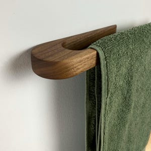 Pode incluir: Um suporte de toalha de madeira com um design curvo. O suporte é feito de madeira de noz e tem um acabamento natural. Uma toalha verde está pendurada no suporte.