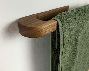 Puede incluir: Un toallero de madera con un diseño curvo. El toallero está hecho de madera de nogal y tiene un acabado natural. Una toalla verde está colgada en el toallero.