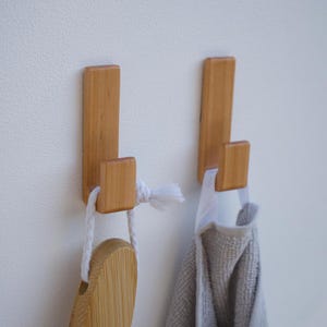 Peut inclure: Deux patères murales en bois clair montées verticalement. Le crochet de gauche soutient une brosse en bois avec une boucle en corde blanche, tandis que celui de droite porte une serviette en tissu gris texturé avec une boucle blanche.