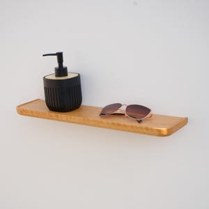 Puede incluir: Una estantería de madera clara con un borde frontal curvado. En la estantería hay un dispensador de jabón negro con una tapa de madera y un par de gafas de sol con montura de oro rosa y lentes marrones. La estantería está montada en una pared blanca.