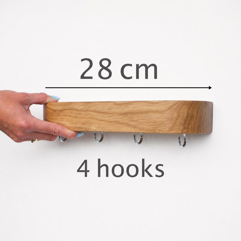 May include: A light brown wooden wall-mounted rack featuring four silver-colored metal hooks along the bottom edge. The rack has a rounded rectangular shape and a visible wood grain texture. The text 28 cm and 4 hooks is displayed.