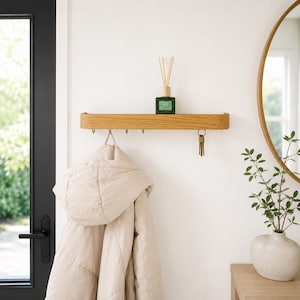 May include: A light oak wood wall-mounted shelf featuring a flat top surface and five metal hooks underneath. The shelf holds a small green glass bottle with reed diffusers, while a set of metal keys hangs from one of the hooks below the shelf.
