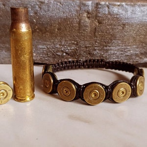 Op de afbeelding: Een zwarte paracord armband met vijf gouden kogelhulzen. De armband ligt op een wit oppervlak naast twee gouden kogelhulzen en een gouden kogelhülse.