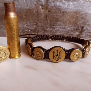 Può includere: Braccialetto fatto a mano con bossoli in ottone e un emblema centrale. Il braccialetto è realizzato con un cordoncino intrecciato marrone scuro. Sono presenti anche due bossoli in ottone e un proiettile.