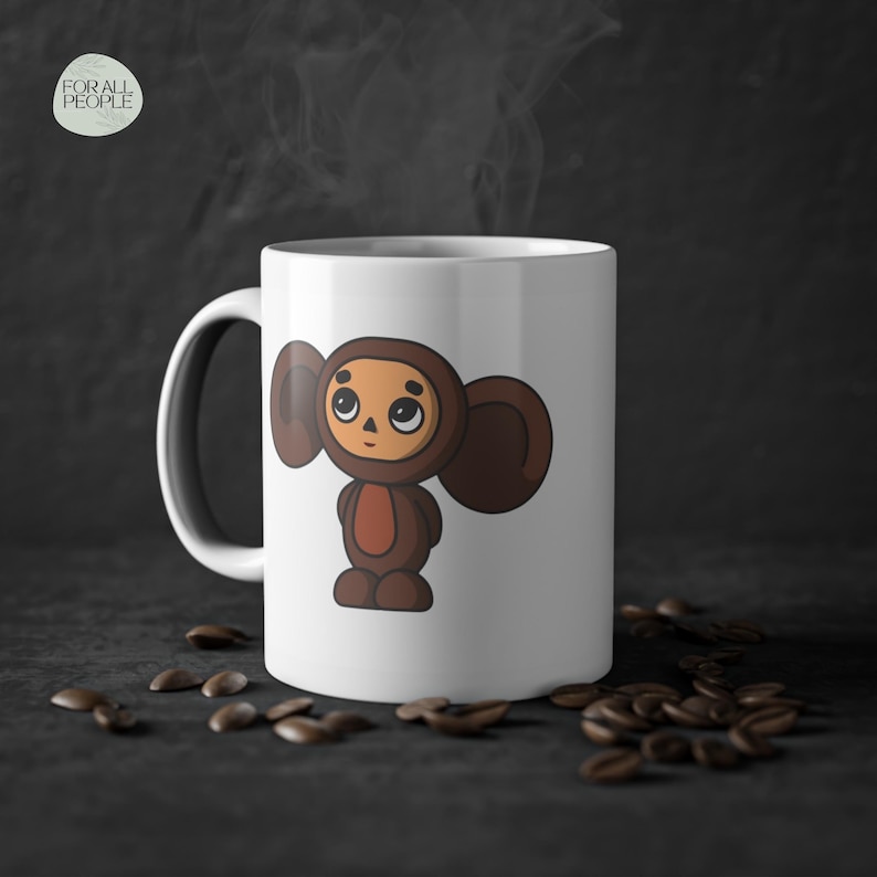 May include: White ceramic mug with a cartoon illustration of a brown character with large ears. The character has a round face with brown eyes and a small mouth. The mug is sitting on a dark surface with coffee beans scattered around it.