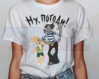 May include: A white short-sleeve t-shirt featuring a graphic print of the cartoon characters Wolf and Hare from the series Nu, Pogodi!. The wolf wears a blue and white striped shirt and sailor hat, while the hare wears a green shirt and shorts.