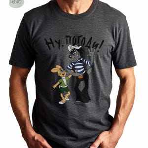 May include: A dark grey t-shirt with a cartoon image of a wolf and a rabbit. The wolf is wearing a sailor hat and striped shirt. The text "Ну, погоди!" is printed on the shirt.