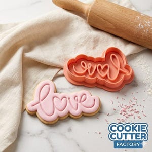 May include: A peach-coloured plastic cookie cutter shaped like the word love in cursive script, positioned next to a matching light pink frosted cookie shaped like the word love, with a wooden rolling pin visible in the background.
