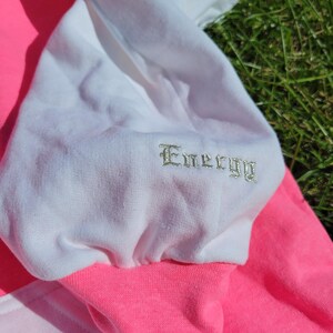 May include: Close-up of a colour-blocked sweatshirt. The sweatshirt features a white sleeve with the word "Energy" embroidered in a silver thread. The cuff and body are bright pink, with a lighter pink panel visible.