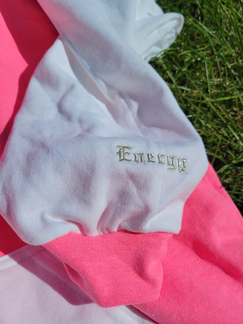 May include: Close-up of a colour-blocked sweatshirt. The sweatshirt features a white sleeve with the word "Energy" embroidered in a silver thread. The cuff and body are bright pink, with a lighter pink panel visible.