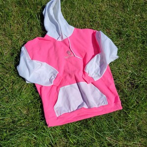 May include: A vibrant pink hoodie with white sleeves, hood, and pocket. The hoodie features a drawstring and a small embroidered design on the chest. The garment is laid on green grass.