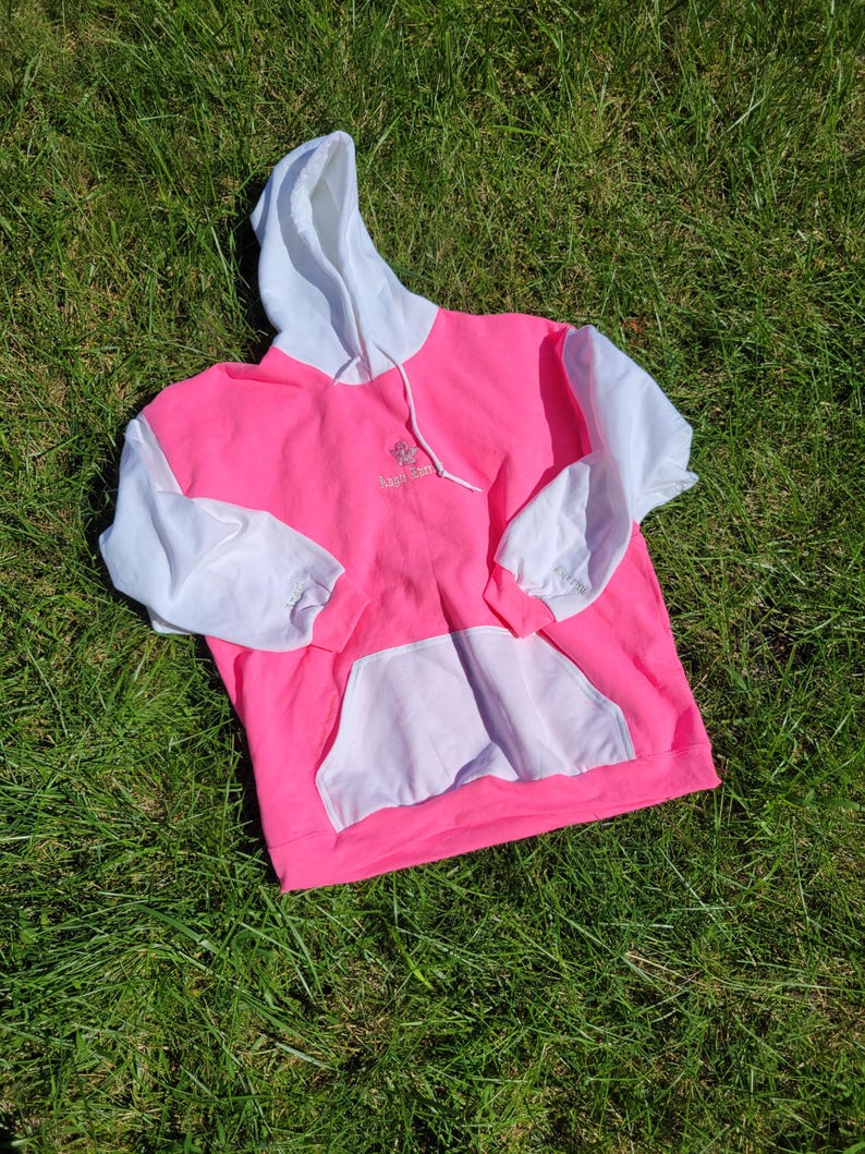 May include: A vibrant pink hoodie with white sleeves, hood, and pocket. The hoodie features a drawstring and a small embroidered design on the chest. The garment is laid on green grass.
