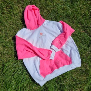 May include: White and pink hoodie with a kangaroo pocket. The sleeves and hood are bright pink, while the body is white. The text "Sage Escape" is embroidered on the chest. The hoodie is lying on a bed of green grass.