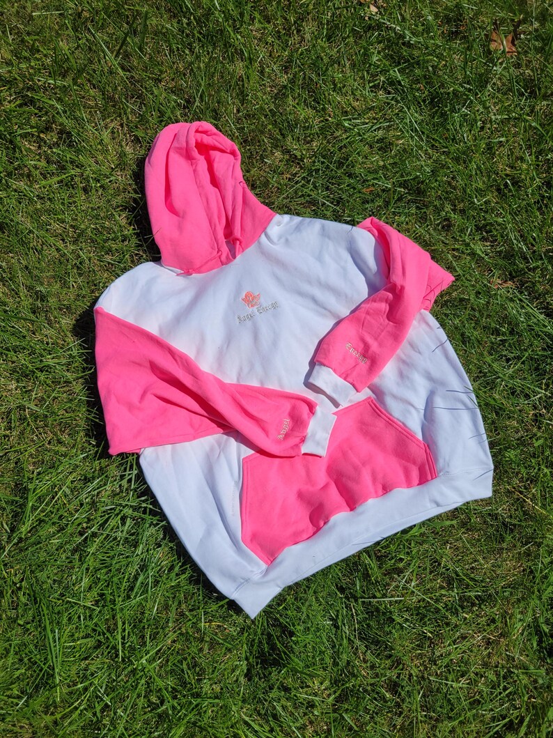 May include: White and pink hoodie with a kangaroo pocket. The sleeves and hood are bright pink, while the body is white. The text "Sage Escape" is embroidered on the chest. The hoodie is lying on a bed of green grass.