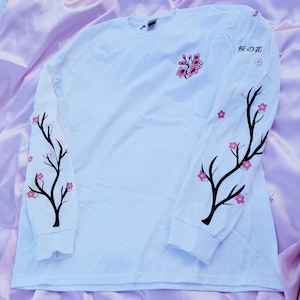 May include: White long-sleeve t-shirt with black branches and pink cherry blossoms printed on the sleeves. The text "桜の花" is printed on the right sleeve.