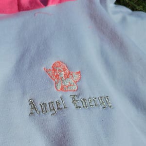 May include: White hoodie with a bright pink hood and drawstring. Features an embroidered cherub design and the words "Angel Energy" in silver thread. A casual, comfortable garment.