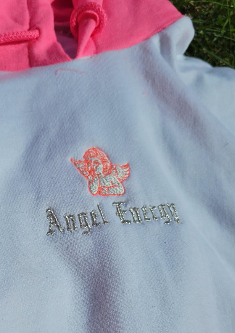 May include: White hoodie with a bright pink hood and drawstring. Features an embroidered cherub design and the words "Angel Energy" in silver thread. A casual, comfortable garment.
