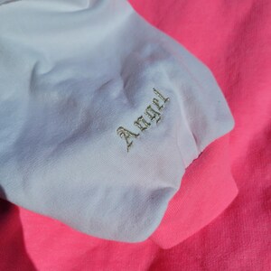 May include: Close-up of a pink and white sweatshirt sleeve. The white sleeve has the word "Angel" embroidered in gold thread. The cuff and body of the sweatshirt are bright pink.