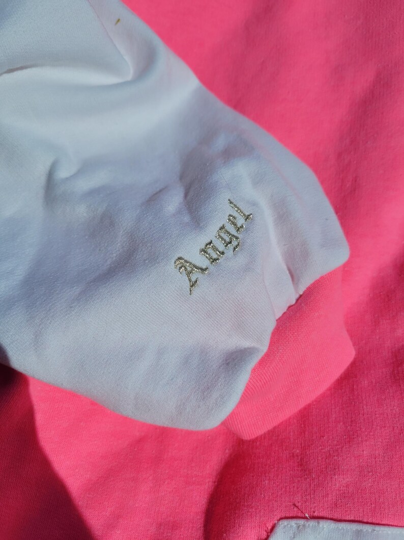 May include: Close-up of a pink and white sweatshirt sleeve. The white sleeve has the word "Angel" embroidered in gold thread. The cuff and body of the sweatshirt are bright pink.