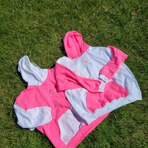 May include: Two white and pink hoodies are laid out on green grass. The hoodies feature a white body, pink sleeves, and a pink hood. Each hoodie has a kangaroo pocket and a small logo on the chest.