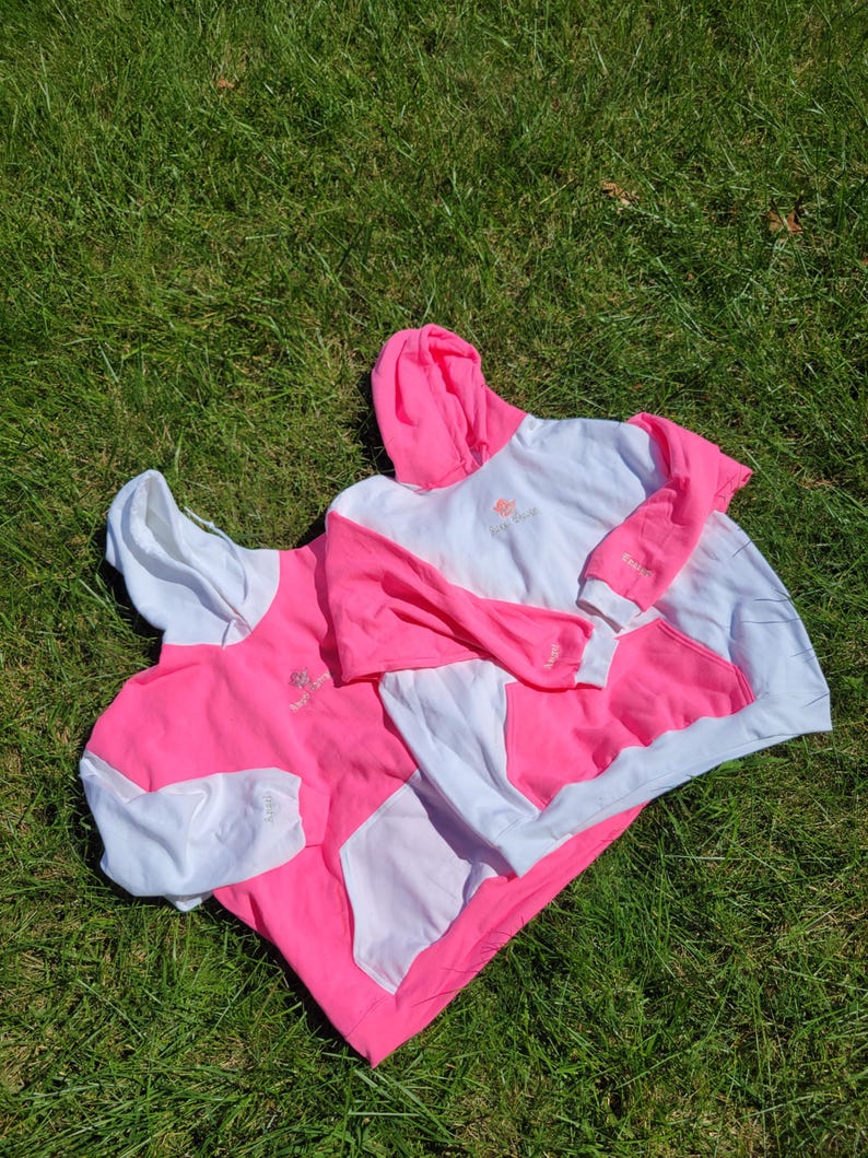 May include: Two white and pink hoodies are laid out on green grass. The hoodies feature a white body, pink sleeves, and a pink hood. Each hoodie has a kangaroo pocket and a small logo on the chest.