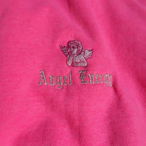May include: Bright pink sweatshirt with a white collar. It has an embroidered cherub and the words "Angel Energy" in silver thread. The material looks soft and comfortable, suitable for casual wear.