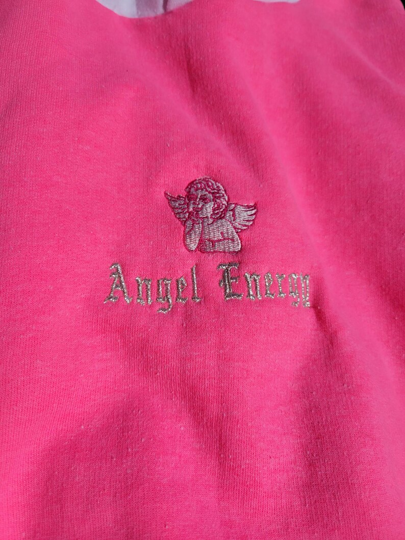 May include: Bright pink sweatshirt with a white collar. It has an embroidered cherub and the words "Angel Energy" in silver thread. The material looks soft and comfortable, suitable for casual wear.
