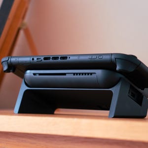 May include: A black handheld gaming console rests on a black charging dock. The console has buttons and ports along the top edge. The dock has a rectangular base with a slot for the console to sit in. The setup is on a wooden surface.
