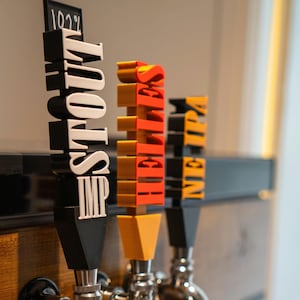 May include: Three chrome beer taps with black handles and colorful labels. The labels read "IMP STOUT", "HELLES", and "NE IPA".