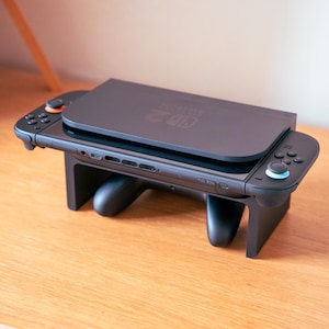 May include: A black Nintendo Switch console with attached controllers and a charging dock. The console is resting on a black stand, with the controllers on either side. The stand is on a wooden surface.