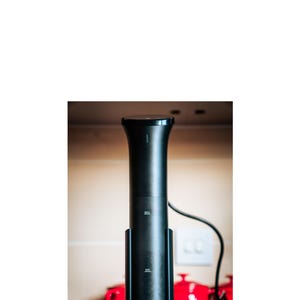 May include: A black immersion circulator with a cylindrical design, featuring 'MAX' and 'MIN' level indicators. The device is set on a dark surface, with red teapots in the background. A black power cord extends from the top.