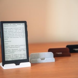 May include: A black e-reader device displaying text on its screen, positioned upright in a white plastic stand. Three additional stands in light grey, brown, and black are arranged in a row behind the device on a flat surface.