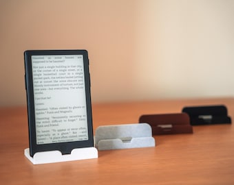 May include: A black e-reader device displaying text on its screen, positioned upright in a white plastic stand. Three additional stands in light gray, brown, and black are arranged in a row behind the device on a flat surface.