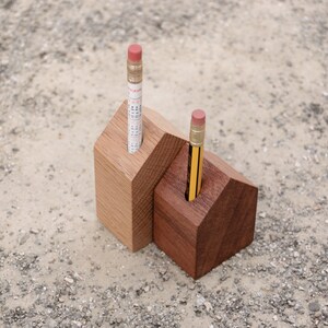 Könnte beinhalten: Zwei stiftförmige Holzstifthalter in Hausform. Einer ist hell, der andere dunkel. Jeder hält einen Bleistift mit einem rosa Radiergummi. Ein Bleistift hat einen aufgedruckten Kalender. Die Halter stehen auf einer strukturierten Oberfläche.