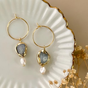 Può includere: Orecchini a cerchio dorati con pietra azzurra e perla bianca. Gli orecchini sono presentati su un piatto bianco con bordo dorato, con fiori secchi sullo sfondo.